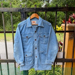 May include: Light blue denim jacket with four front pockets and brown buttons. The jacket has a collared neckline and long sleeves. A small red tag is visible on the front pocket. The jacket is hanging on a wooden hanger.