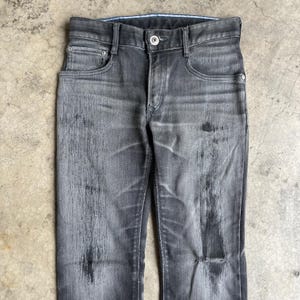 May include: A pair of faded black denim jeans. The jeans have a classic five-pocket design with a button and zipper closure. The denim has a worn appearance with distressed areas and subtle variations in color.