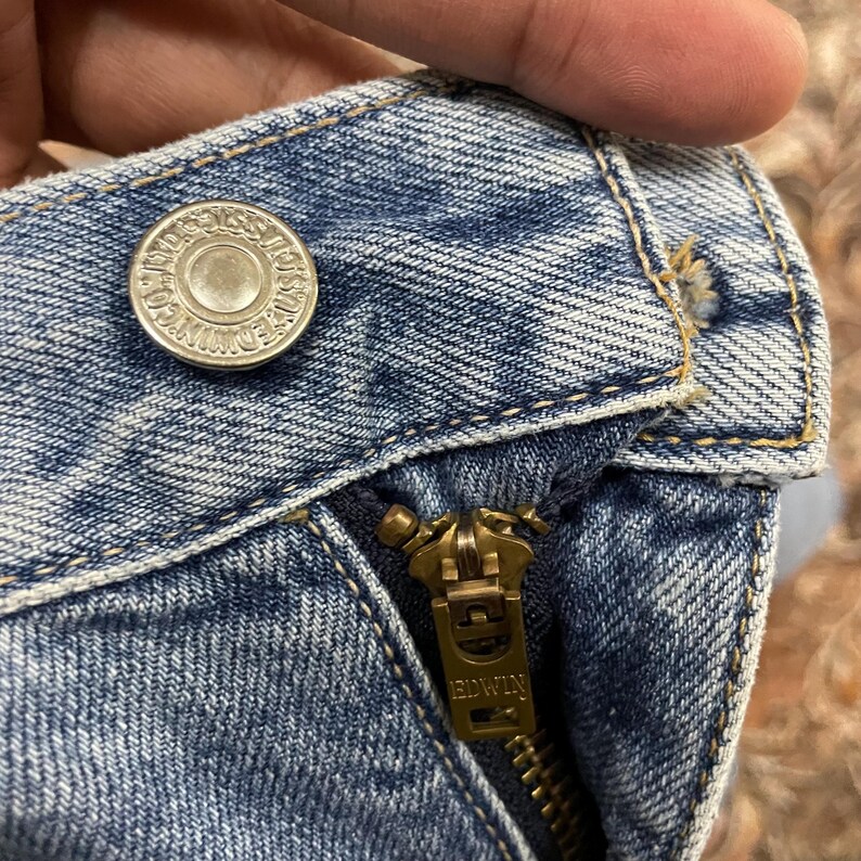 May include: Light blue acid-wash denim jeans featuring a silver-toned metal button embossed with the text EDWIN CO. LTD. and a brass-toned metal zipper pull tab clearly engraved with the brand name EDWIN, showcasing visible gold-colored stitching.