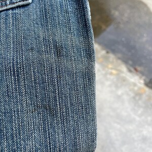 May include: Close-up of blue denim fabric, showing the texture of the weave and the stitching details. The denim has a classic, worn appearance with subtle variations in color and some darker spots.