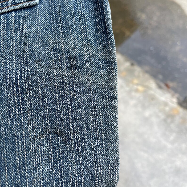 May include: Close-up of blue denim fabric, showing the texture of the weave and the stitching details. The denim has a classic, worn appearance with subtle variations in color and some darker spots.