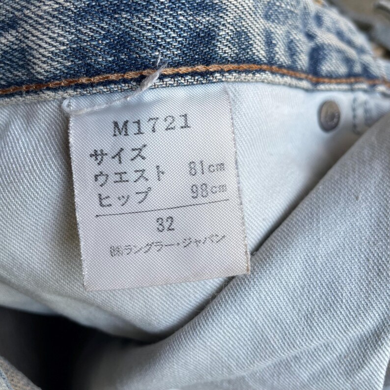 May include: Close-up of a light blue denim jeans label. The label displays the product code M1721, waist size 81cm, hip size 98cm, and size 32. The label also includes Japanese text.