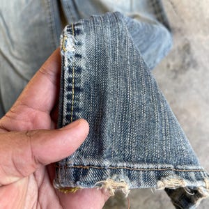 May include: Close-up of light blue denim jeans. The jeans show wear and tear, with frayed edges and visible stitching. The fabric has a textured appearance, and the overall look is casual and worn.
