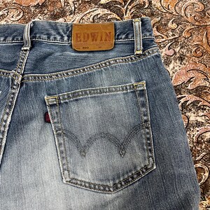 May include: Blue denim jeans featuring a tan leather patch on the back waistband with the text EDWIN 503. The jeans have a single back pocket with a curved stitched design and a small red fabric tab on the side seam, showcasing a faded wash.