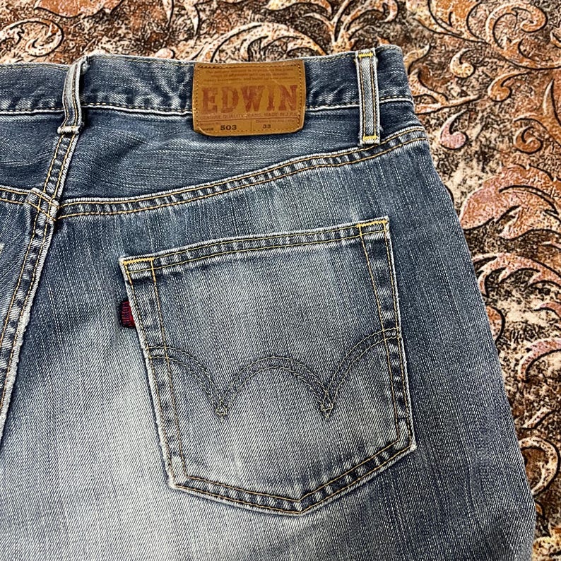 May include: Blue denim jeans featuring a tan leather patch on the back waistband with the text EDWIN 503. The jeans have a single back pocket with a curved stitched design and a small red fabric tab on the side seam, showcasing a faded wash.