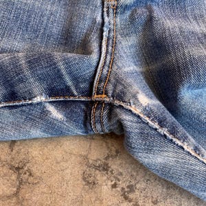 Pu&ograve; includere: Primo piano della zona del cavallo di jeans blu usurati. I jeans mostrano segni di usura, con strappi e sbiaditure visibili. Le cuciture sono di un colore arancione-marrone a contrasto.