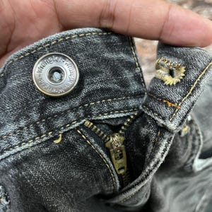 May include: Close-up of the top of a pair of black denim jeans. The image shows the button, zipper, and stitching details. The button is metallic with the word "Wrangler" around the edge.