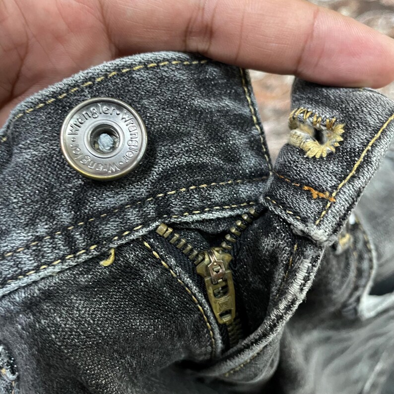 May include: Close-up of the top of a pair of black denim jeans. The image shows the button, zipper, and stitching details. The button is metallic with the word "Wrangler" around the edge.