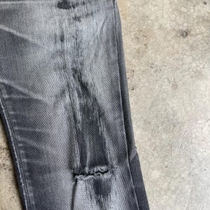 May include: Close-up of distressed gray denim jeans. The jeans have a faded appearance with intentional wear and tear, including a large rip near the knee. The fabric shows vertical streaks and variations in color, creating a worn, vintage aesthetic.