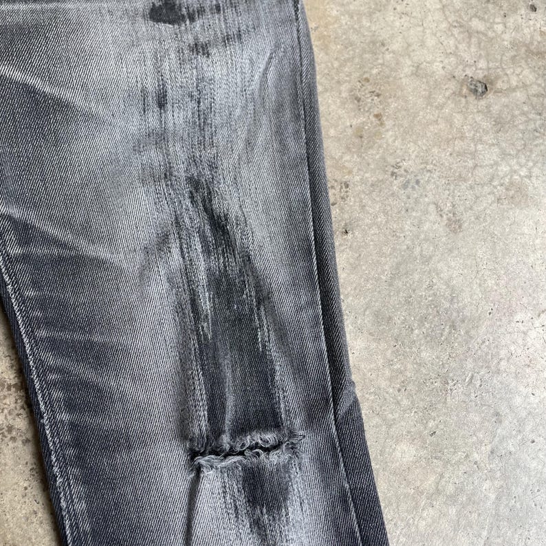 May include: Close-up of distressed gray denim jeans. The jeans have a faded appearance with intentional wear and tear, including a large rip near the knee. The fabric shows vertical streaks and variations in color, creating a worn, vintage aesthetic.