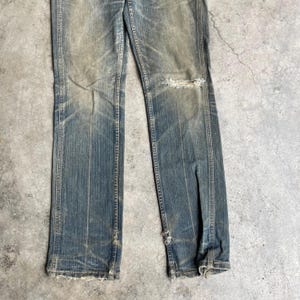May include: Faded blue denim jeans with distressed details. The jeans have a straight leg cut, with a ripped section on the right knee and frayed edges at the bottom. The denim shows signs of wear and fading.