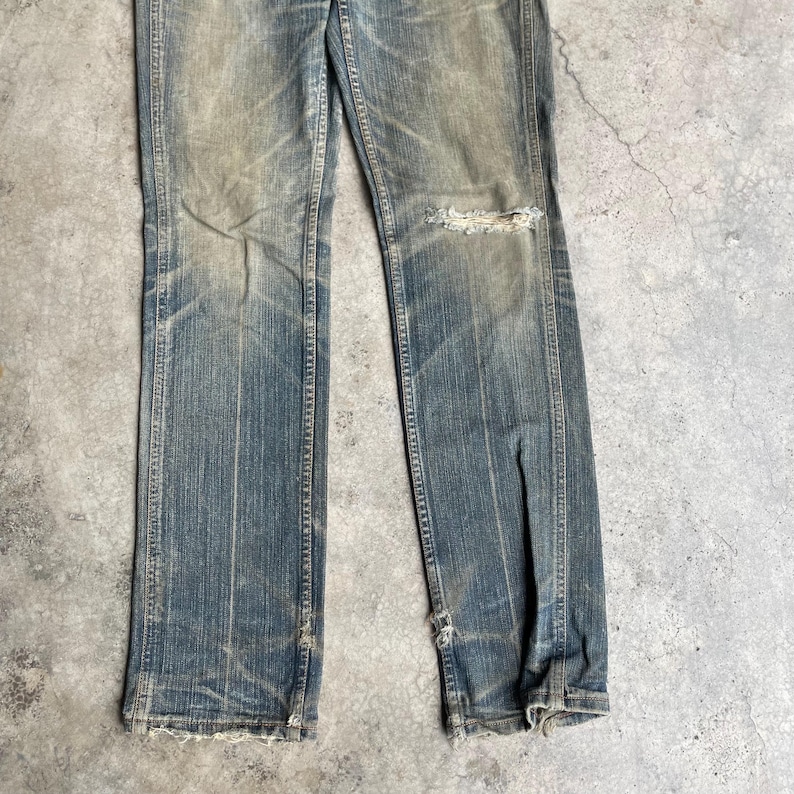 May include: Faded blue denim jeans with distressed details. The jeans have a straight leg cut, with a ripped section on the right knee and frayed edges at the bottom. The denim shows signs of wear and fading.