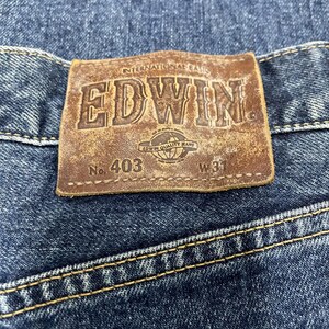 Puede incluir: Primer plano de una etiqueta de cuero marr&oacute;n cosida a unos vaqueros azules. La etiqueta muestra el nombre de la marca "EDWIN" en relieve, junto con el texto "INTERNATIONAL BASIC" y "EDWIN QUALITY BRAND". Los vaqueros est&aacute;n marcados con "No. 403" y "W31".