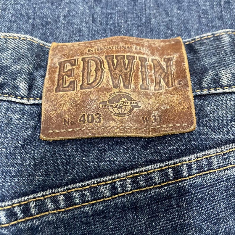 Puede incluir: Primer plano de una etiqueta de cuero marr&oacute;n cosida a unos vaqueros azules. La etiqueta muestra el nombre de la marca "EDWIN" en relieve, junto con el texto "INTERNATIONAL BASIC" y "EDWIN QUALITY BRAND". Los vaqueros est&aacute;n marcados con "No. 403" y "W31".