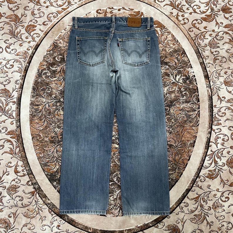 May include: A pair of light blue denim jeans featuring a straight-leg cut, two back pockets with a signature arcuate stitch design, a small red tab on the right pocket, and a tan leather patch on the waistband embossed with the brand name EDWIN.