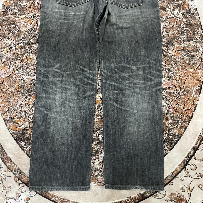 May include: Faded black denim jeans with a straight leg cut. The jeans have a worn appearance with visible fading and distressed detailing. The back pockets and waistband are visible. The jeans are displayed on a patterned surface.