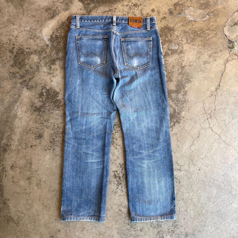 Pu&ograve; includere: Jeans in denim blu chiaro con lavaggio sbiadito e dettagli usurati. La vista posteriore mostra le tasche e una toppa in pelle marrone con il marchio "EDWIN". I jeans hanno un classico design a cinque tasche.