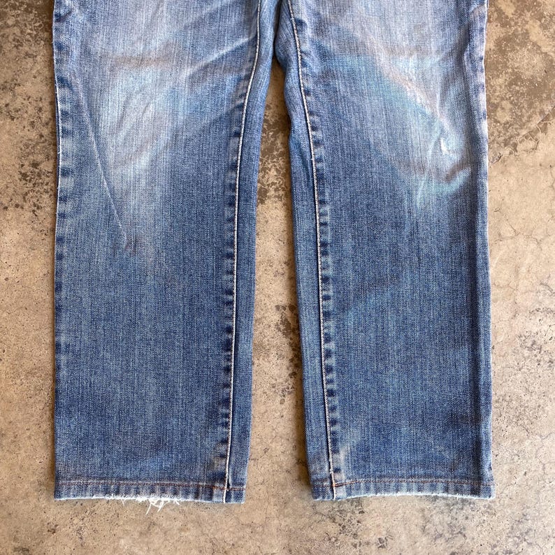 Pu&ograve; includere: Primo piano di un paio di jeans blu chiaro. I jeans hanno un lavaggio sbiadito e cuciture bianche. Gli orli inferiori sono sfilacciati. I jeans sono realizzati in resistente denim.