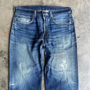 May include: Faded blue denim jeans with a classic five-pocket design. The jeans show signs of wear with distressed areas and a light wash, creating a vintage aesthetic. The jeans have a button fly and belt loops.