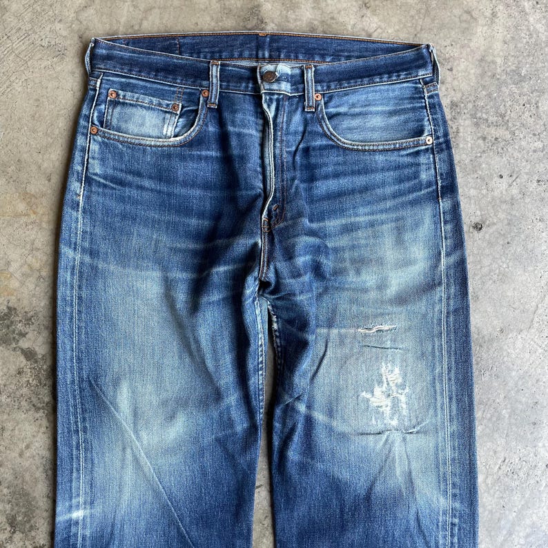 May include: Faded blue denim jeans with a classic five-pocket design. The jeans show signs of wear with distressed areas and a light wash, creating a vintage aesthetic. The jeans have a button fly and belt loops.