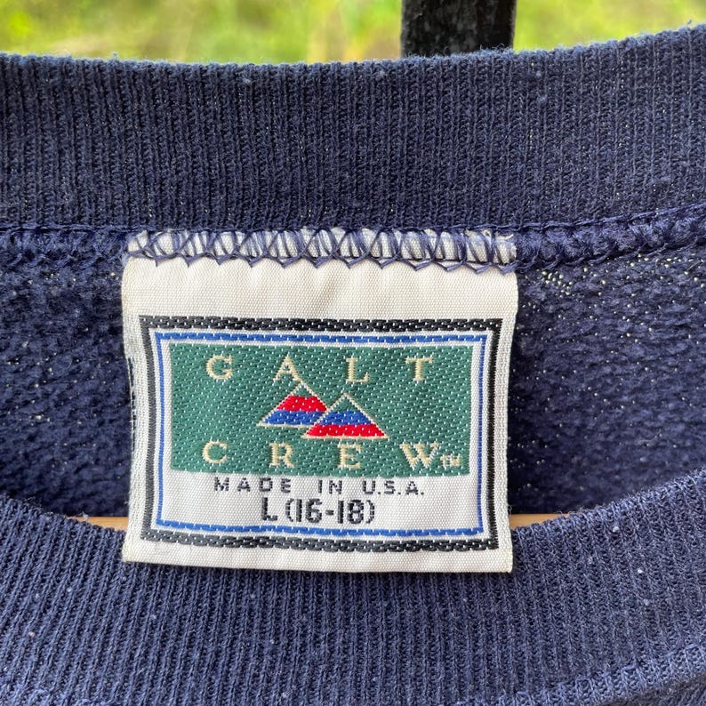 May include: Close-up of a navy blue sweatshirt with a tag that reads "GALT CREW MADE IN U.S.A." The tag features a green rectangle with the brand name and two red and blue mountain-like shapes. The size is L (16-18).