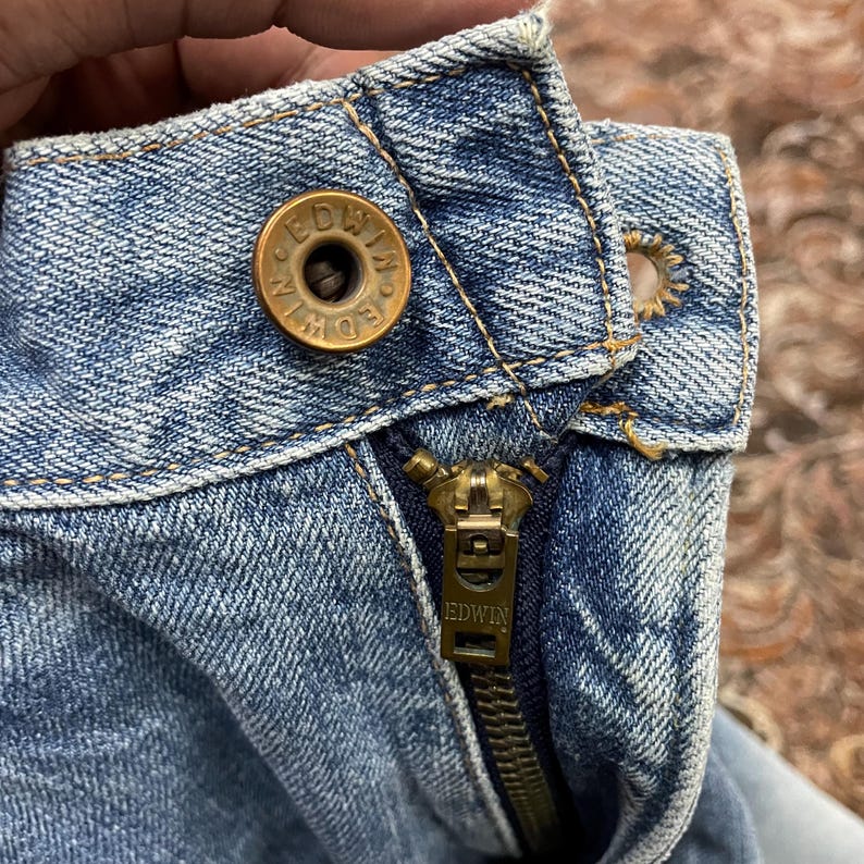 May include: Close-up of a light blue denim garment. The image highlights a brass-colored button with the brand name "EDWIN" and a matching zipper. The zipper is partially open, revealing the denim's texture and stitching.