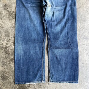 May include: Blue denim jeans with a faded wash. The jeans have a classic five-pocket design, a button and zip closure, and a straight leg. The hem is slightly frayed. The jeans are made of a durable denim fabric.