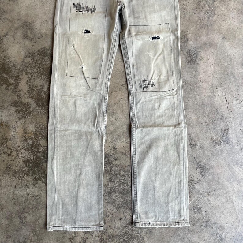 K&ouml;nnte beinhalten: Hellgraue Jeans mit aufgerissenen Patches und dekorativen N&auml;hten. Die Jeans hat einen geraden Beinverlauf und ein verwaschenes Aussehen mit sichtbaren Gebrauchsspuren.