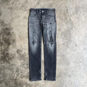 May include: Dark gray denim jeans with a distressed, faded appearance. The jeans have a button and zipper closure, classic five-pocket design, and a ripped knee. The fabric shows wear and tear, adding to the vintage aesthetic.