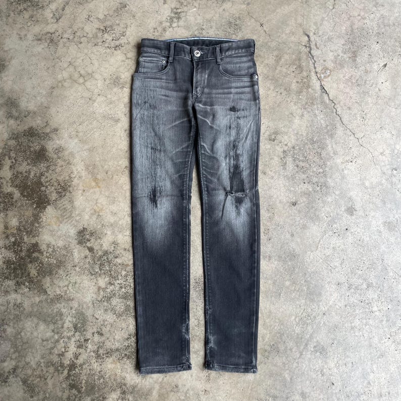 May include: Dark gray denim jeans with a distressed, faded appearance. The jeans have a button and zipper closure, classic five-pocket design, and a ripped knee. The fabric shows wear and tear, adding to the vintage aesthetic.