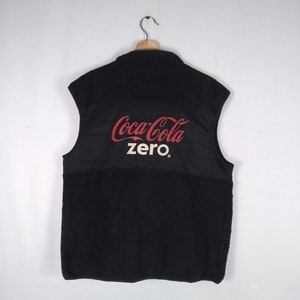 Puede incluir: Chaleco de forro polar negro con un logotipo de Coca-Cola Zero en la espalda. El logotipo es rojo y blanco con las palabras "Coca-Cola" en negrita y "Zero" en una fuente más pequeña.