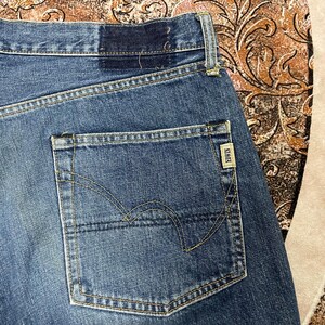 May include: Blue denim jeans featuring a single back pocket with yellow contrast stitching and a small rectangular beige tag with the text EDWIN sewn onto the edge. The waistband includes a dark blue patch detail near the belt loop area.