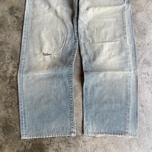 May include: Light blue denim jeans with a distressed, worn appearance. The jeans have a ripped knee and a faded wash. The straight-leg style features visible stitching and a frayed hem, suggesting a vintage or well-loved aesthetic.