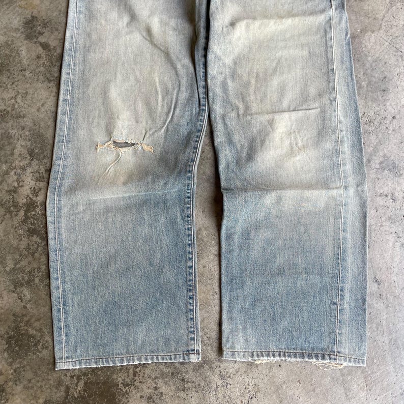 May include: Light blue denim jeans with a distressed, worn appearance. The jeans have a ripped knee and a faded wash. The straight-leg style features visible stitching and a frayed hem, suggesting a vintage or well-loved aesthetic.