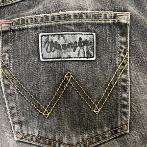 May include: Close-up of a black denim jeans pocket. The pocket features a leather patch with the brand name "Wrangler" and a signature "W" stitching design in yellow thread. The top edge has a pink accent.