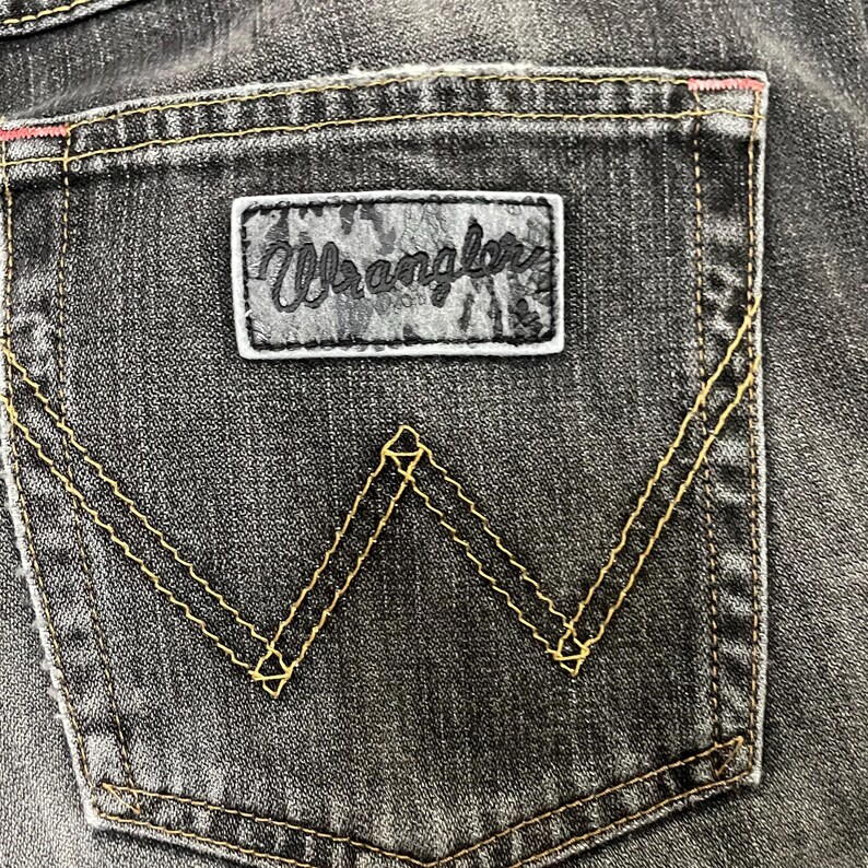 May include: Close-up of a black denim jeans pocket. The pocket features a leather patch with the brand name "Wrangler" and a signature "W" stitching design in yellow thread. The top edge has a pink accent.