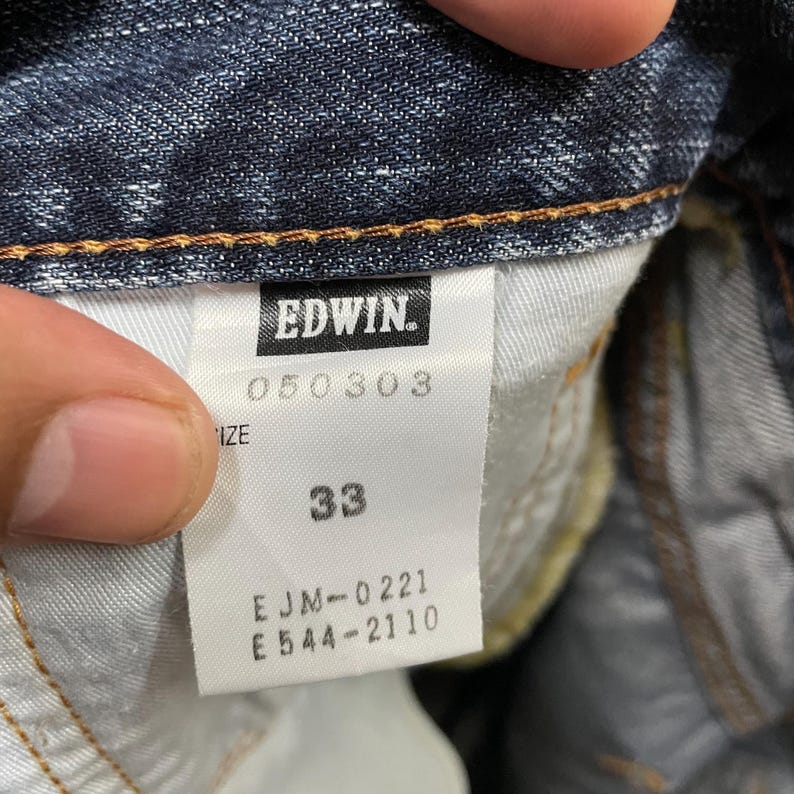 May include: A white interior garment label attached to dark blue denim fabric. The label features the brand name EDWIN in a black box, the number 050303, the size 33, and the alphanumeric codes EJM-0221 and E544-2110 printed in black text.