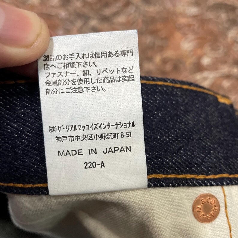 May include: Close-up of a white tag with Japanese text and English text that reads "MADE IN JAPAN" and "220-A". The tag is attached to a dark blue denim garment. A copper-colored button is visible.