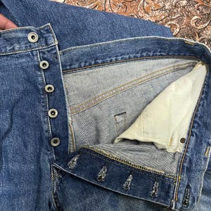 May include: A pair of blue denim jeans featuring a button-fly closure with five silver-toned metal buttons. The interior shows a light beige cotton pocket lining and visible orange contrast stitching along the seams and the waistband construction.
