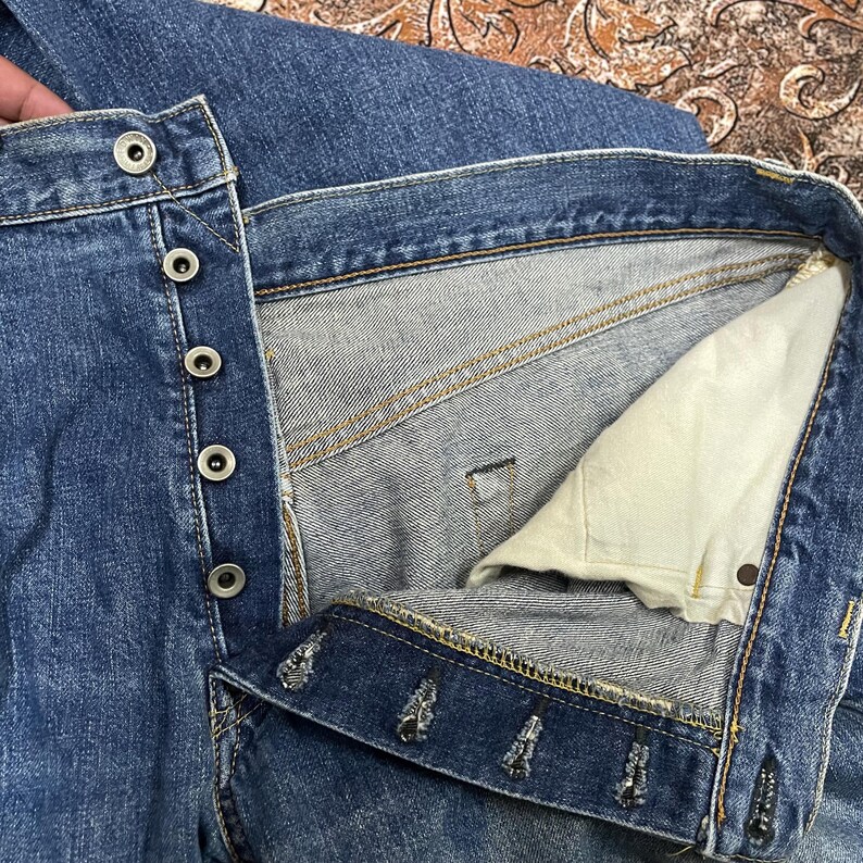 May include: A pair of blue denim jeans featuring a button-fly closure with five silver-toned metal buttons. The interior shows a light beige cotton pocket lining and visible orange contrast stitching along the seams and the waistband construction.