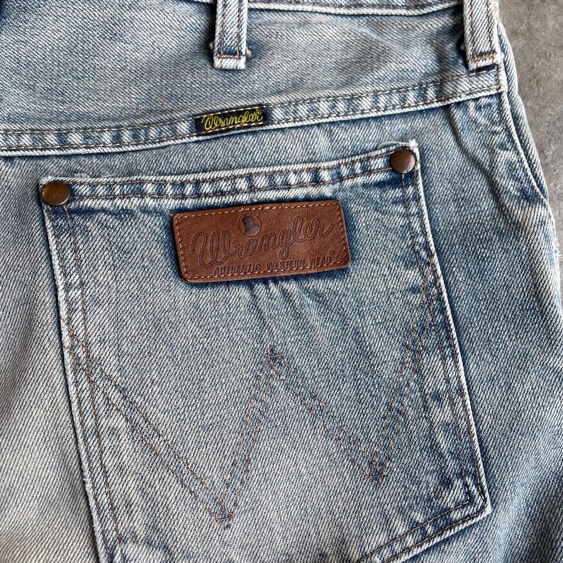 May include: Close-up of light blue denim jeans featuring a back pocket with a brown leather Wrangler patch. The patch has the Wrangler logo and the words "AUTHENTIC WESTERN WEAR". The jeans have a classic five-pocket design.