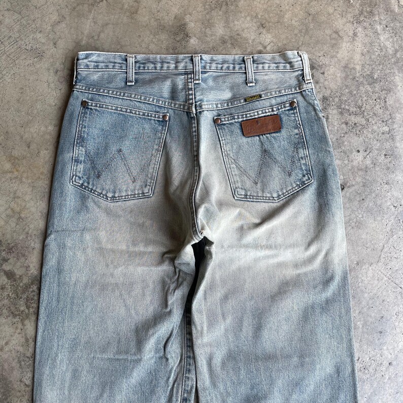 May include: Light blue denim jeans with a faded wash effect. The jeans feature two back pockets with the Wrangler logo and a brown leather patch. The jeans have a classic five-pocket design and a straight leg cut.