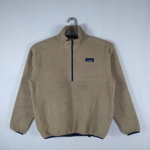 Puede incluir: Un jersey de polar de color beige con cierre de cremallera de media cremallera y un parche con el logotipo de LL Bean de color azul marino en el pecho.