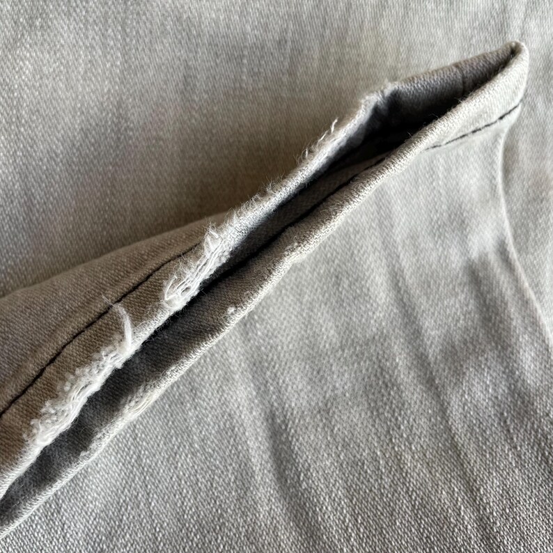 K&ouml;nnte beinhalten: Nahaufnahme von hellgrauem Denim-Stoff, die den gefalteten Saum einer Jeans zeigt. Der Saum ist mit schwarzem Faden gen&auml;ht und hat einen ausgefransten Rand, der die Textur und die Konstruktionsdetails des Stoffes zeigt.