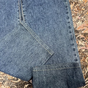 Puede incluir: Primer plano de unos vaqueros azules. Los vaqueros tienen un dise&ntilde;o cl&aacute;sico de cinco bolsillos, con cremallera y cierre de bot&oacute;n. Las costuras est&aacute;n reforzadas con pespuntes amarillos. El denim tiene un lavado medio y un aspecto ligeramente descolorido.