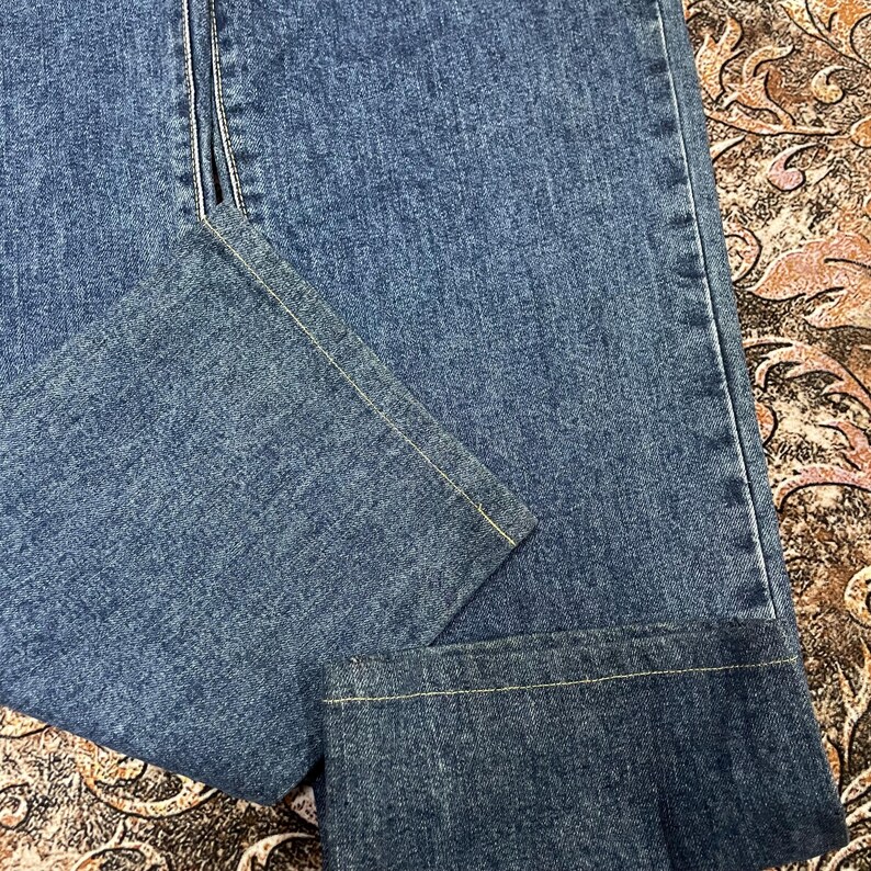 Puede incluir: Primer plano de unos vaqueros azules. Los vaqueros tienen un dise&ntilde;o cl&aacute;sico de cinco bolsillos, con cremallera y cierre de bot&oacute;n. Las costuras est&aacute;n reforzadas con pespuntes amarillos. El denim tiene un lavado medio y un aspecto ligeramente descolorido.