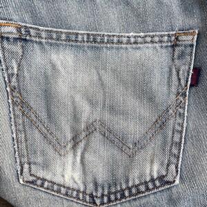 May include: Close-up of a light blue denim jeans pocket. The pocket features a classic design with a stitched 'W' shape and brown stitching. A small red tag is visible on the right side of the pocket.