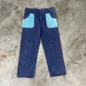 May include: Dark blue pants with two large, light blue front pockets. The pockets are outlined with a contrasting stitch. The pants have a button closure and a straight leg design. The fabric appears to be a sturdy cotton or similar material.
