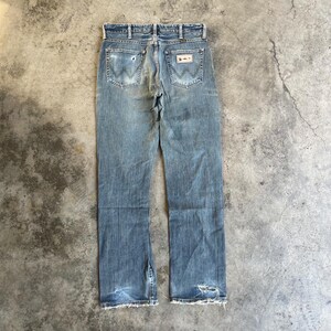 May include: Faded blue denim jeans with a classic five-pocket design. The jeans feature the Wrangler logo on the back pockets and show signs of wear, including distressed areas and frayed edges. The jeans are laid flat on a concrete surface.