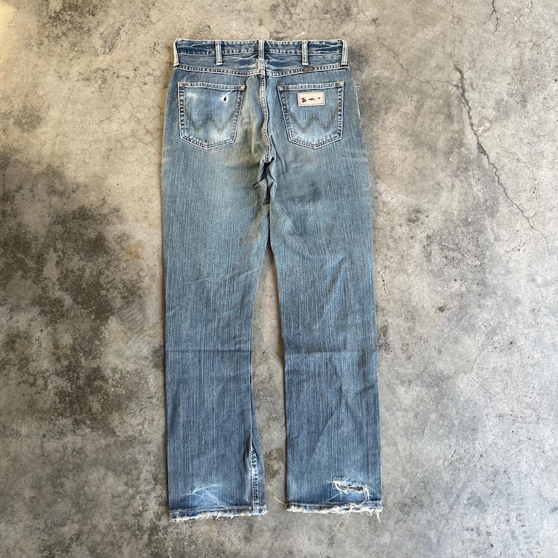 May include: Faded blue denim jeans with a classic five-pocket design. The jeans feature the Wrangler logo on the back pockets and show signs of wear, including distressed areas and frayed edges. The jeans are laid flat on a concrete surface.